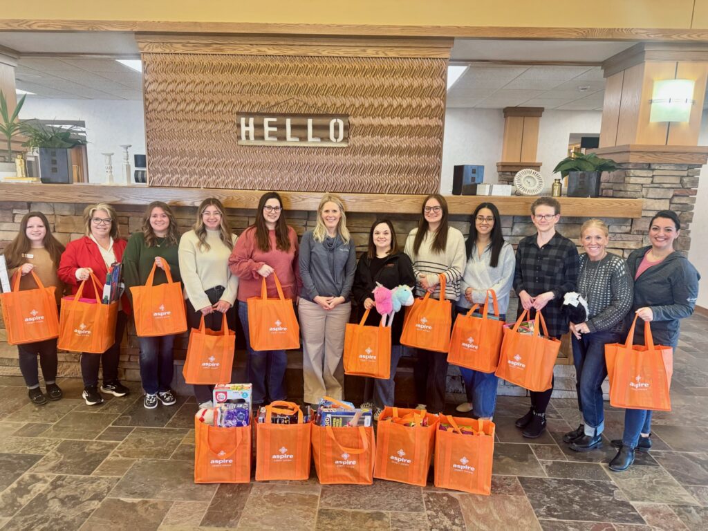 Make A Wish Donation 2026 - Aspire Credit Union Smiling volunteers stand in a lobby holding orange Aspire tote bags, with a decorative wall behind them and a 'HELLO' sign above.